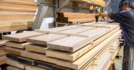 A craftsman holding a moisture meter to a stickered stack of hardwood boards in Wood Depot's timber store, the spacers between planks letting air circulate as the wood acclimatises to Singapore's humidity.
